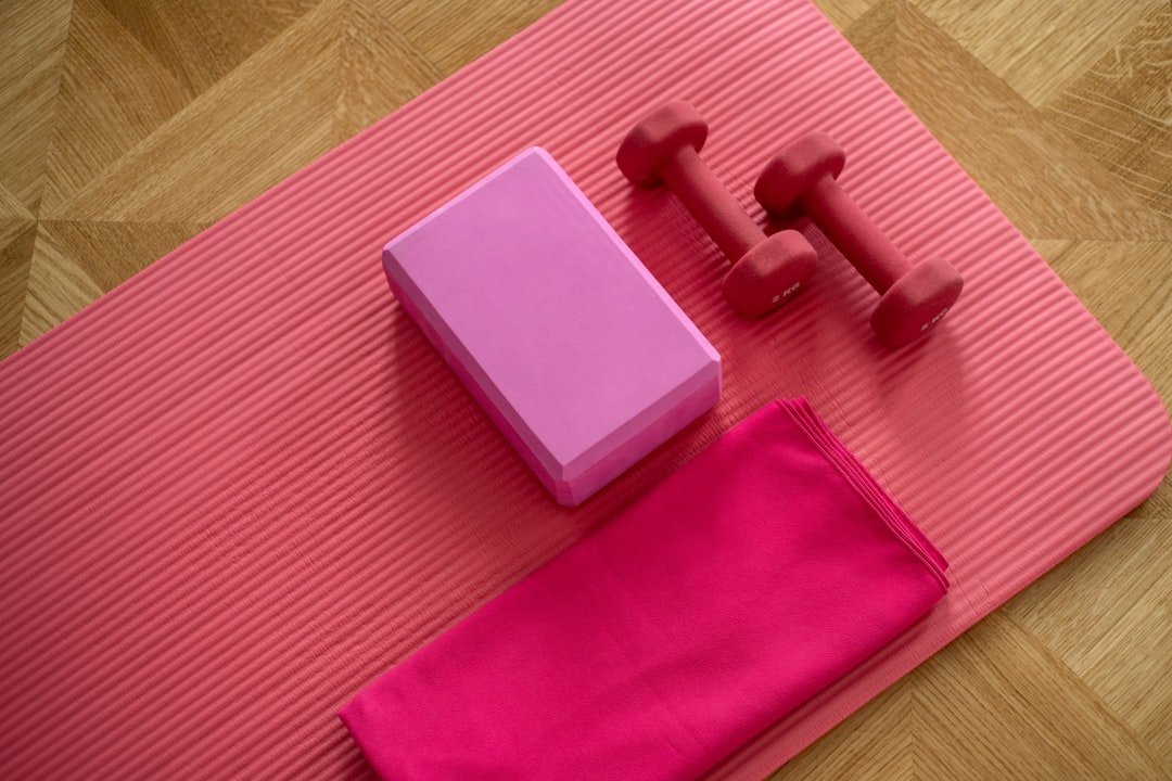 Pink exercise mat, yoga blocks, resistance band, and dumbbells arranged on a wood floor.