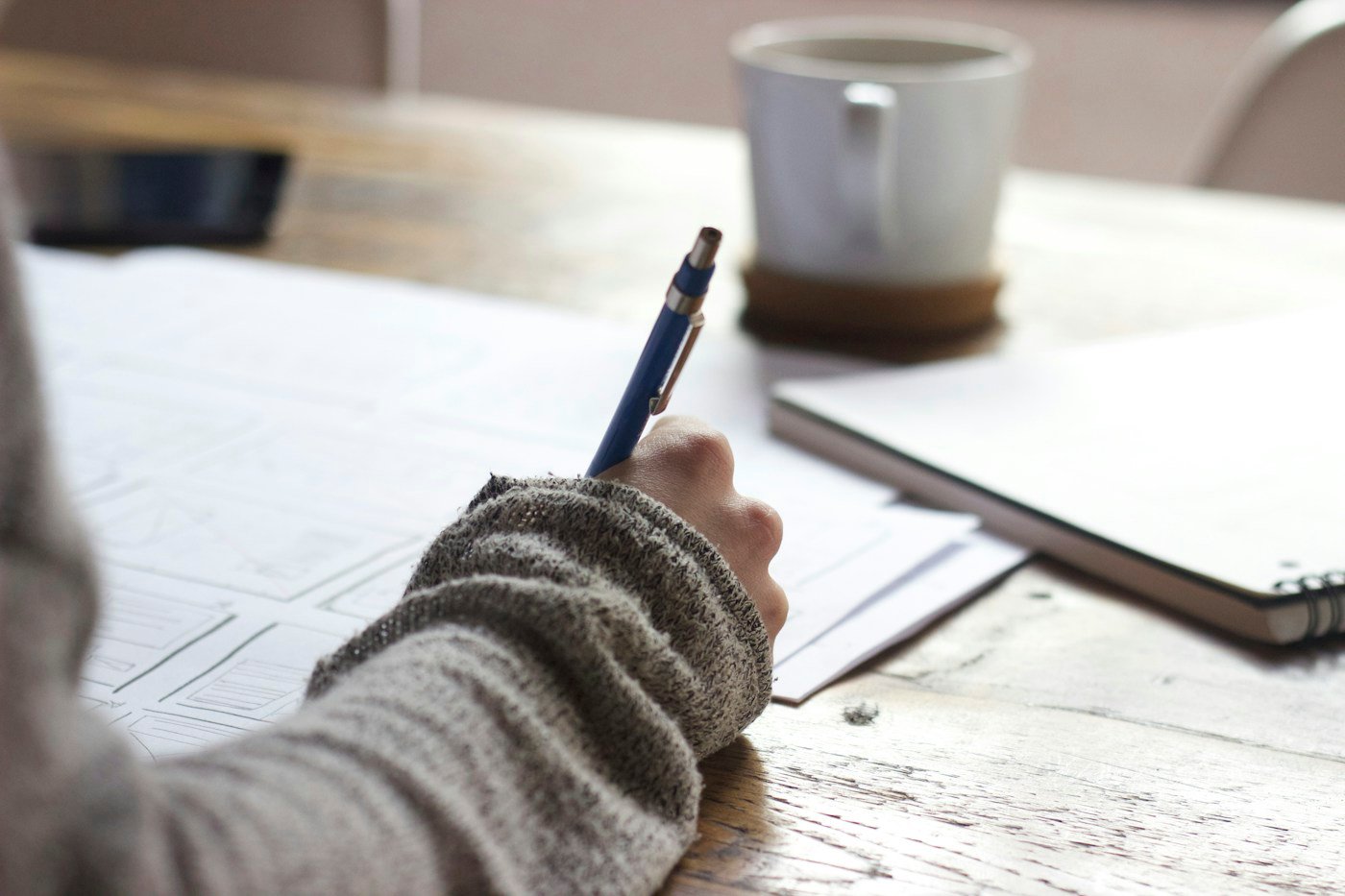 A person writing in a notebook beside a mug at a wooden table.